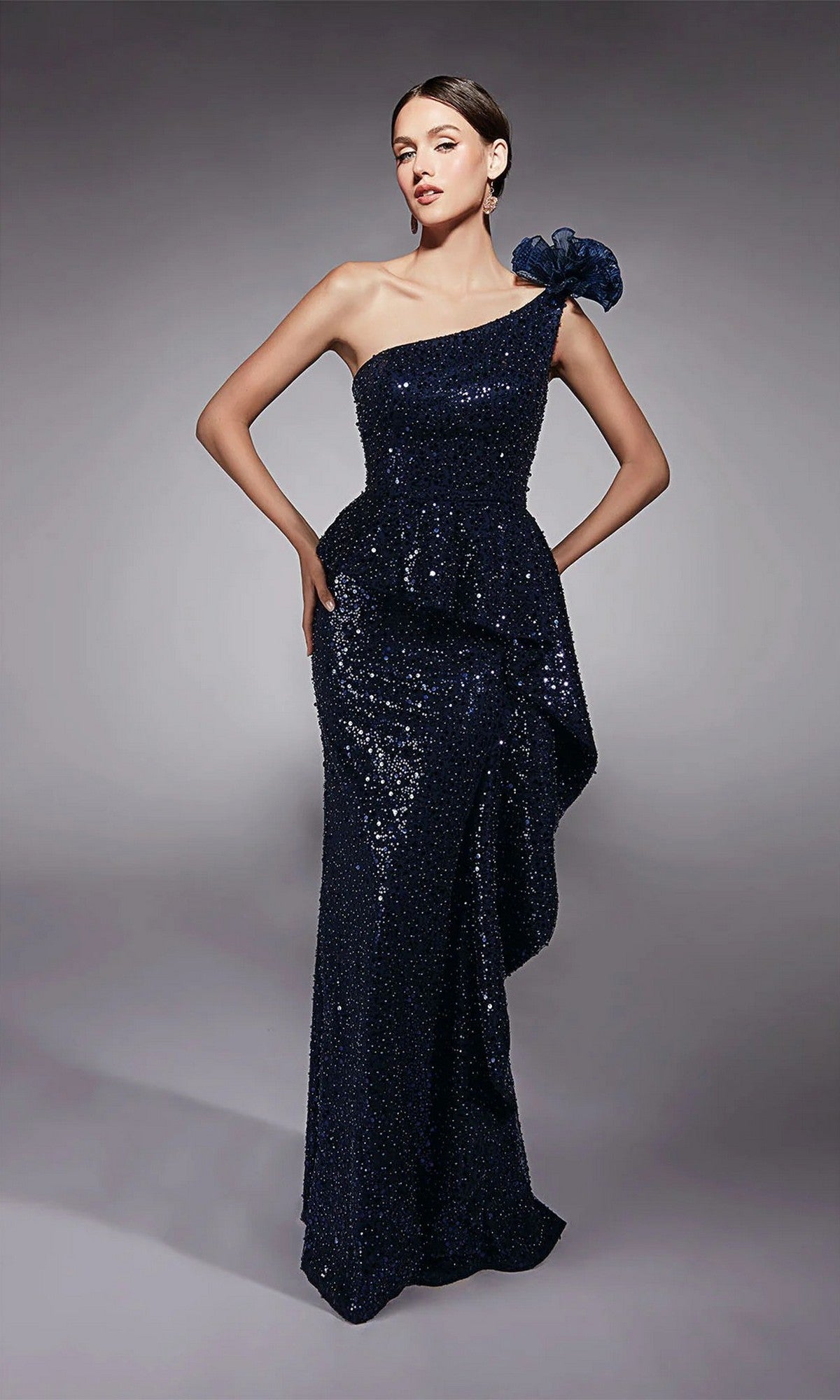 A woman poses against a gray background in the Alyce 27770 long formal dress in navy, featuring dazzling beaded sequins, a one-shoulder floor-length silhouette, ruffled waist detail, and a decorative shoulder bow—ideal as a mother of the bride gown.